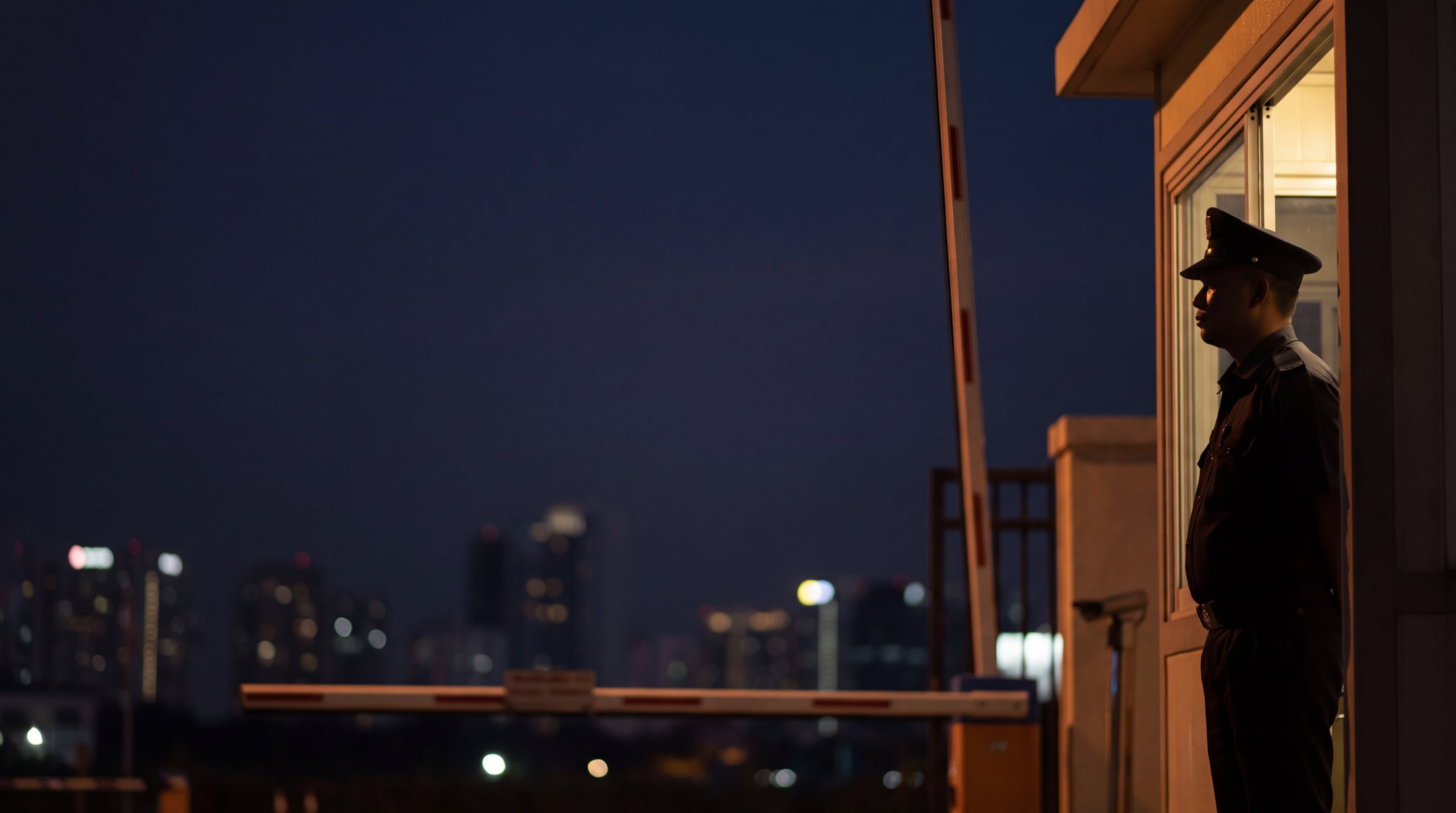 A security officer at a lit guardhouse at night, the city behind